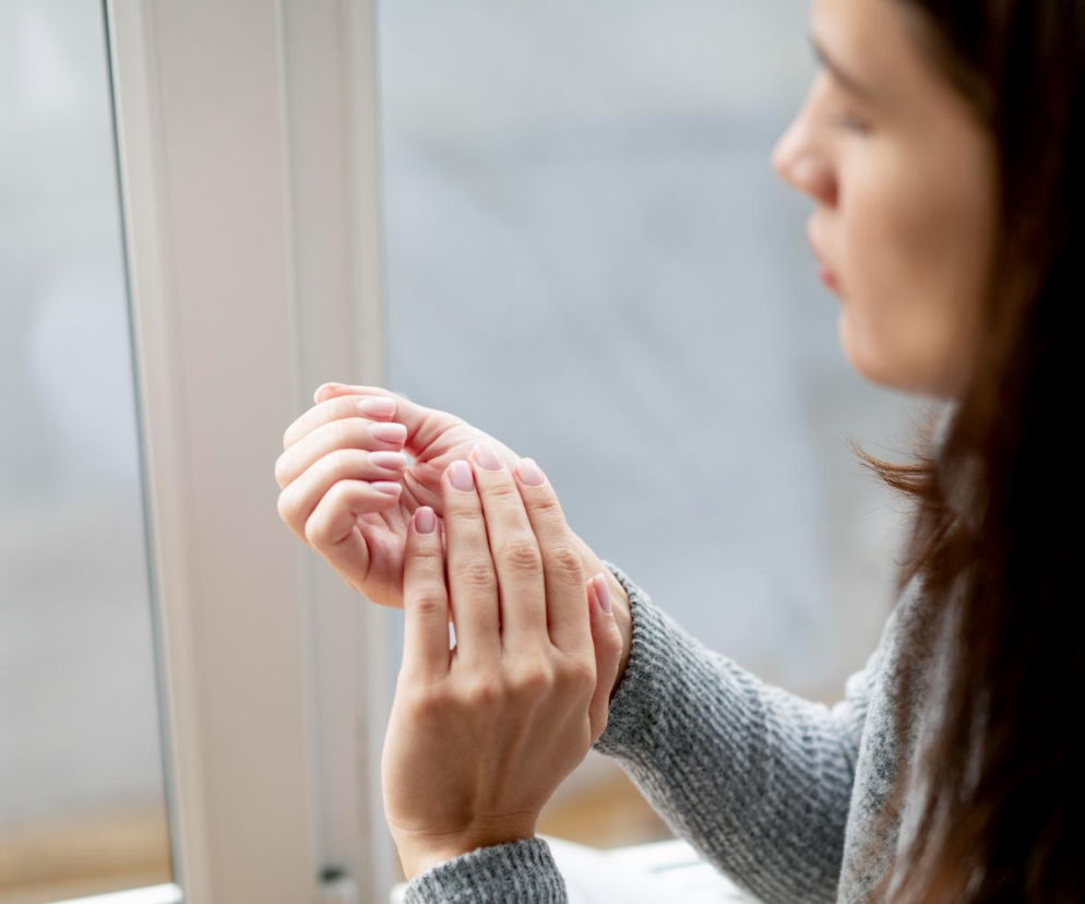 Młoda kobieta w szarym swetrze trzyma jedną dłoń na drugiej, sprawdzając stan skóry rąk, widoczne są zadbane paznokcie z delikatnym manicure. Zdjęcie ilustruje problem suchych dłoni i potrzebę nawilżania, o czym szerzej przeczytasz na Poradnik Zdrowie.