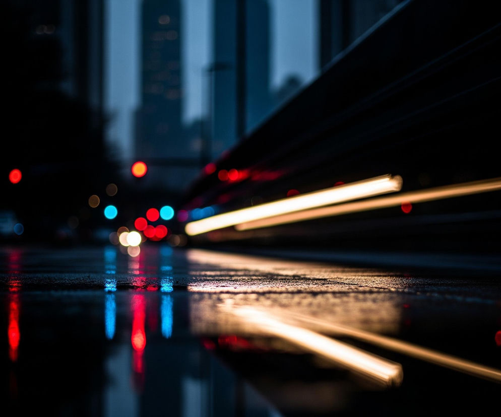 Ulica pokryta jest kroplami wody, które odbijają światła nocnego miasta. Na horyzoncie widoczne są rozmyte światła w kolorach czerwonym, niebieskim i żółtym. Z prawej strony obrazu biegną pod kątem dynamiczne, smużące się paski światła w odcieniach złota i bieli, sugerujące ruch pojazdu. W tle, częściowo widoczne są zarysy budynków w szaro-niebieskich barwach, również rozmazane, co dodatkowo podkreśla efekt prędkości i nocnej scenerii.