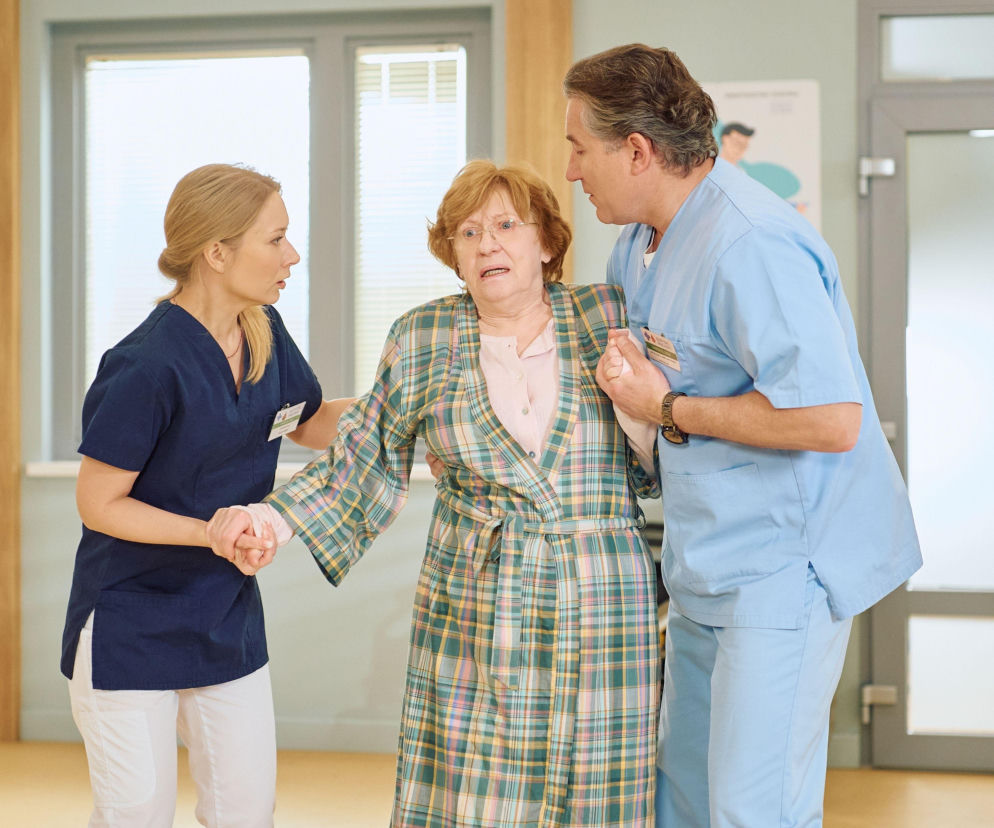 Dwóch pracowników służby zdrowia w jasnych uniformach, kobieta i mężczyzna, wspierają starszą pacjentkę w kraciastej szacie, która wydaje się zaniepokojona. Scena z serialu Na Wspólnej, gdzie Agierowa zmaga się z problemami zdrowotnymi, a widzowie mogą śledzić rozwój wydarzeń na Super Seriale.