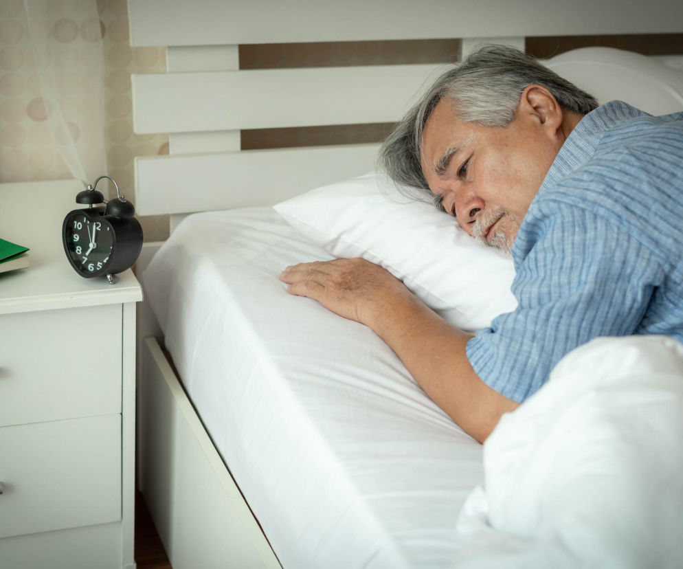Starszy mężczyzna leży w łóżku z otwartymi oczami, nie mogąc zasnąć, obok łóżka budzik wskazujący godzinę 23:40. Ilustracja problemów ze snem przy IBS, o których przeczytasz na Poradnik Zdrowie.