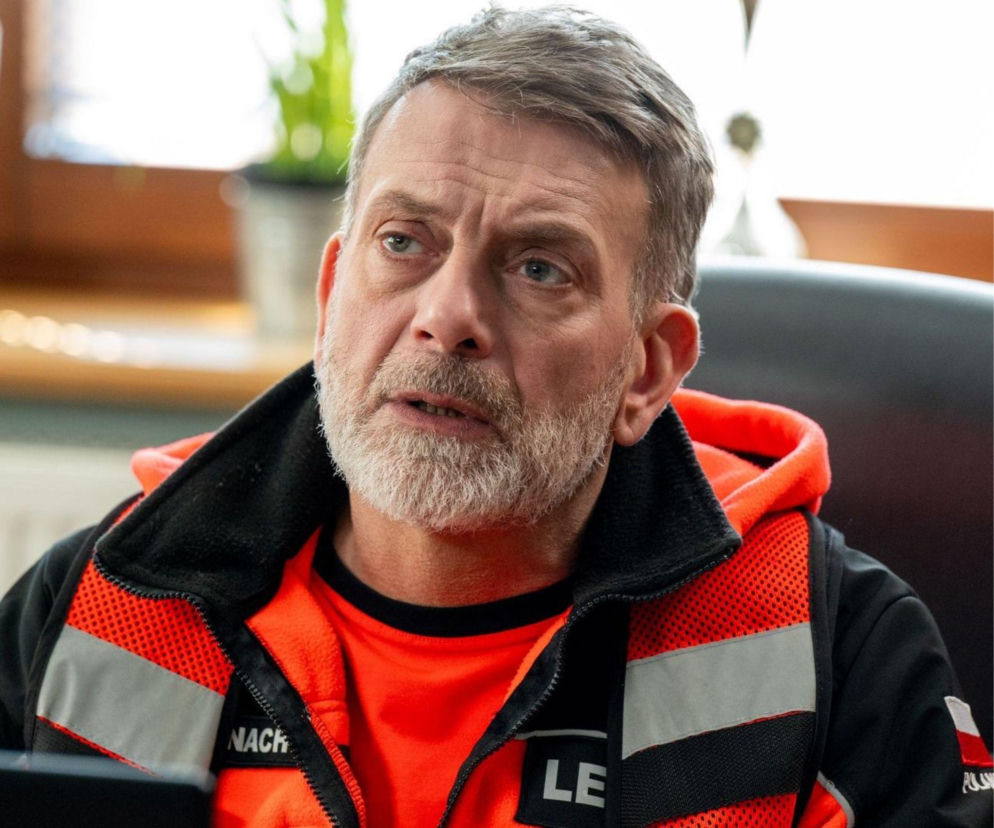 Mężczyzna z siwą brodą i szarymi włosami, ubrany w pomarańczową bluzę i czarno-pomarańczową kurtkę ratownika medycznego z odblaskowymi pasami. Jego twarz wyraża zaniepokojenie, a usta są lekko otwarte, jakby właśnie coś mówił. Tło jest rozmyte, co skupia uwagę na postaci, która prawdopodobnie jest doktorem Góra lub doktorem Banachem z serialu Na sygnale. Więcej informacji o serialu znajdziesz na Super Seriale.