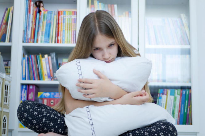 Smutna dziewczynka mocno ściska białą poduszkę na tle regału z książkami. O lęku dziecka w szkole przeczytasz na MJM.