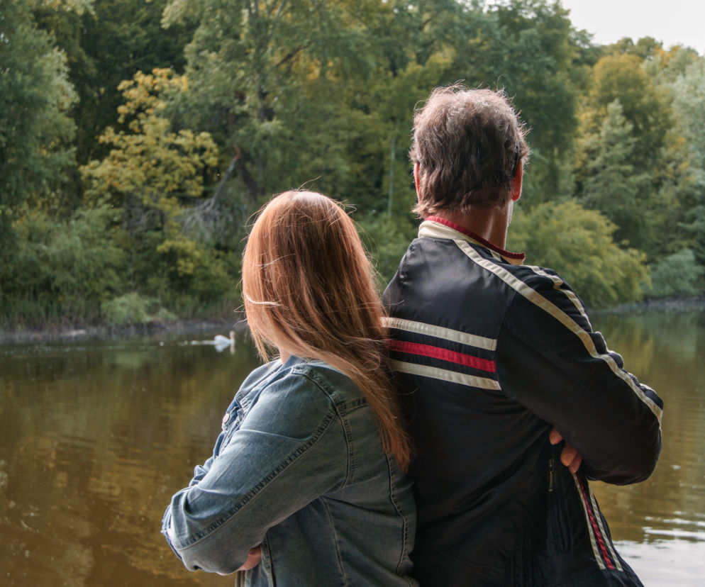 Para, kobieta z rudymi włosami i mężczyzna, stojący tyłem nad jeziorem, z zalesionym brzegiem w tle. Ta fotografia stockowa symbolizuje dylematy Dnia Ojca po rozwodzie, o których możesz przeczytać więcej na naszym portalu.