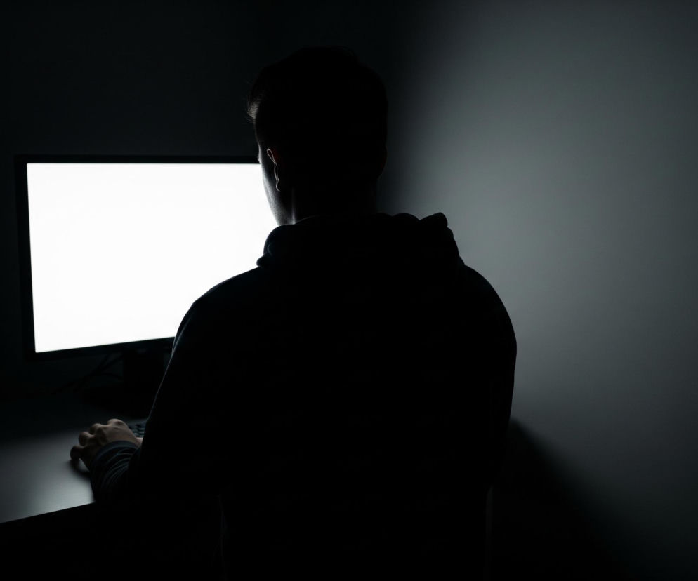 Mężczyzna siedzi tyłem do widza, zwrócony w stronę monitora komputerowego, którego ekran intensywnie świeci bielą. Postać jest całkowicie w cieniu, zarysowana ciemną sylwetką na tle jasno oświetlonego ekranu, z niewielkim odblaskiem widocznym na prawym uchu i karku. Ciemne ubranie, prawdopodobnie bluza z kapturem, z łatwością wtapia się w otoczenie. Po prawej stronie zdjęcia znajduje się ściana w odcieniach ciemnej szarości, podczas gdy lewa strona i dolna część obrazu toną w całkowitej czerni, tworząc głęboki kontrast z białym ekranem. Na biurku, również w ciemnych barwach, widać dłoń mężczyzny spoczywającą na klawiaturze lub myszy, ledwo widoczną w cieniu.