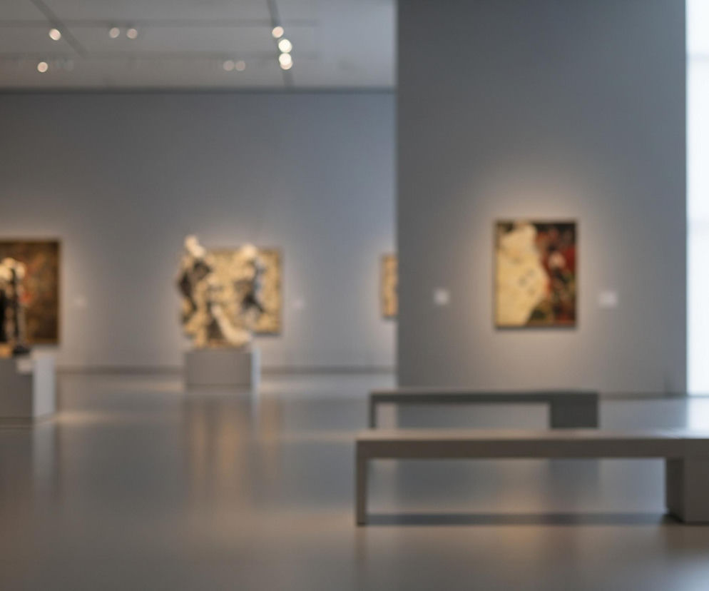Wnętrze galerii sztuki przedstawia ściany w odcieniach szarości i jasną podłogę odbijającą światło. Na ścianach w tle wiszą rozmyte obrazy w ramkach, a w centrum widoczna jest niewyraźna rzeźba na postumencie. Dwa długie, proste ławki w kolorze jasnoszarym, z widocznym cieniem, znajdują się na pierwszym planie po prawej stronie, zachęcając do odpoczynku. W prawym górnym rogu obrazu rozciąga się duża, jasna przestrzeń, prawdopodobnie okno, wpuszczające naturalne światło do pomieszczenia.
