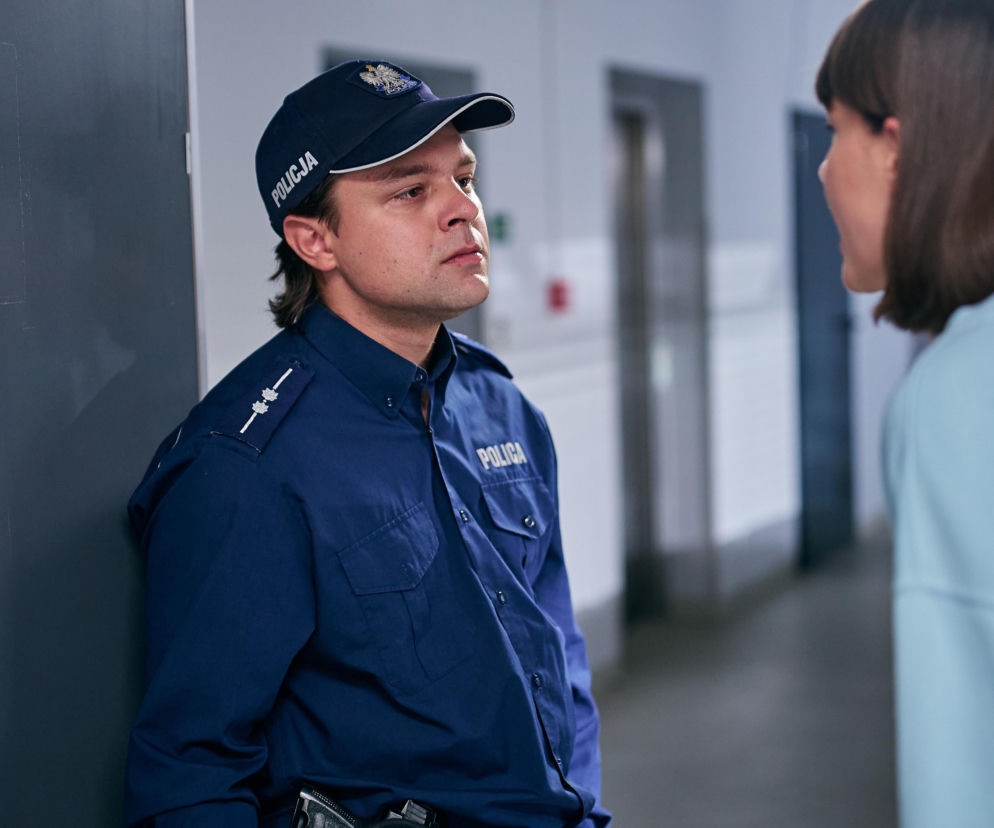 Policjant Klusek w mundurze i czapce policyjnej rozmawia z kobietą w jasnej bluzce. Zdjęcie przedstawia dramatyczne wydarzenia z serialu Na Wspólnej, gdzie Klusek traci wzrok po wypadku na służbie. Więcej o losach bohaterów przeczytasz na Super Seriale.