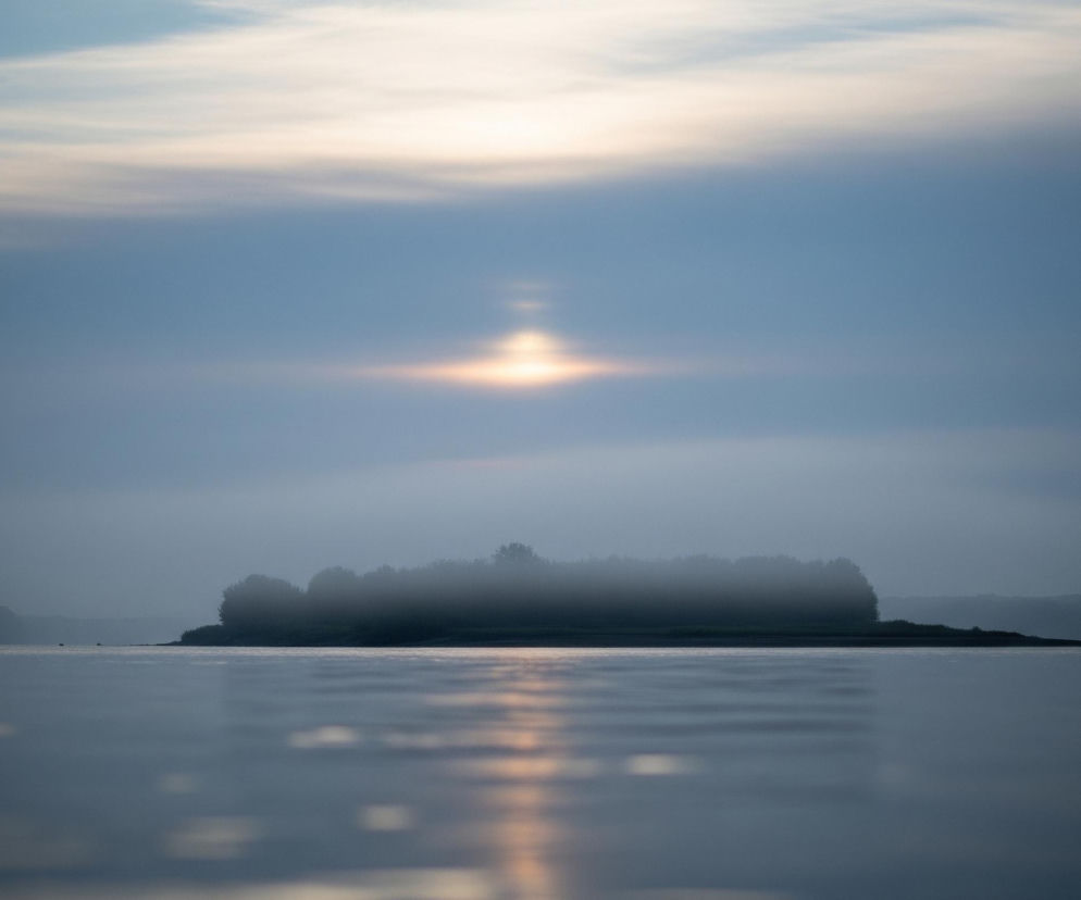 Poniżej linii horyzontu rozciąga się ciemnoniebieska, gładka tafla wody z kilkoma delikatnymi zmarszczkami odbijającymi jasne pasma światła. Na środku obrazu, tuż nad wodą, znajduje się podłużna wyspa pokryta gęstym lasem, częściowo spowita mgłą, która zakrywa jej górne partie. Po obu stronach wyspy widać odległe, zamglone zalesione wzniesienia, blednące w oddali. Ponad horyzontem niebo jest wypełnione warstwami chmur o odcieniach błękitu i szarości, z jasnym, poziomym rozbłyskiem światła słonecznego na środku, przebijającym się przez obłoki i tworzącym pionowe słupy światła.