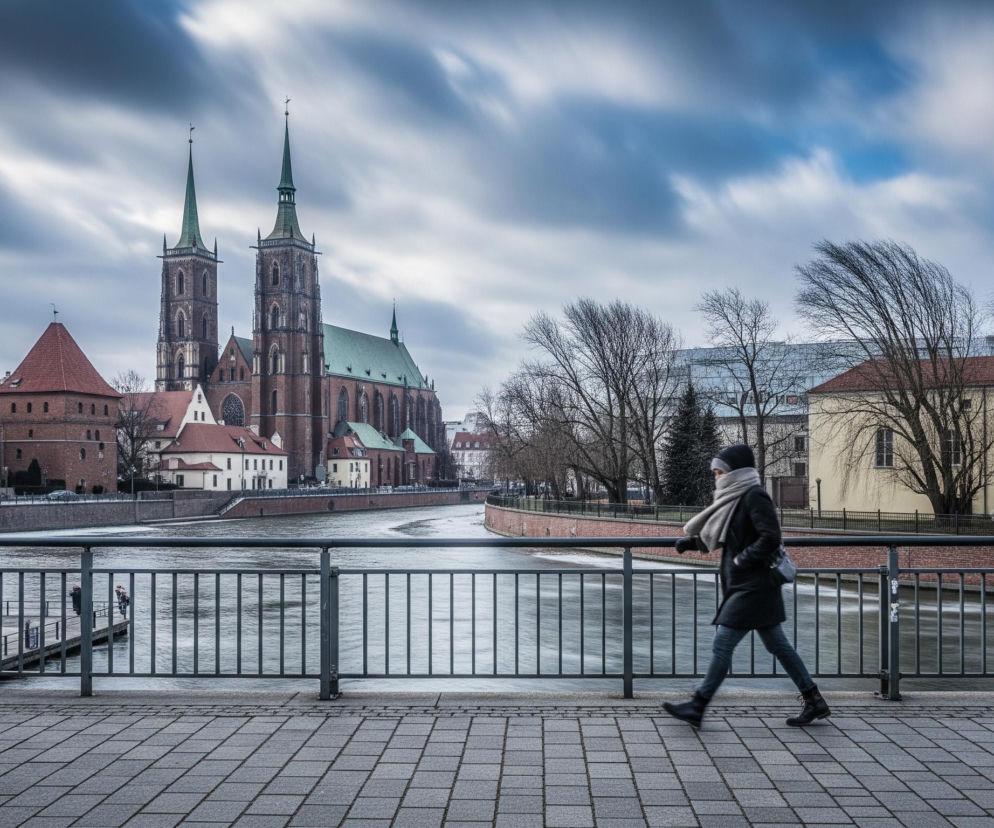 Osoba w ciemnym płaszczu, szarej czapce i szaliku idzie w prawo, wzdłuż chodnika wybrukowanego szarymi prostokątnymi płytami, przed metalową balustradą mostu. W tle płynie rzeka, po lewej stronie widać kamienice i ceglaną gotycką katedrę z dwoma wysokimi, spiczastymi wieżami z zielonymi dachami, a po prawej stronie rzeki stoją budynki z czerwonymi dachami i drzewa bez liści. Niebo jest pochmurne, z rozmyciem chmur wskazującym na długi czas ekspozycji.