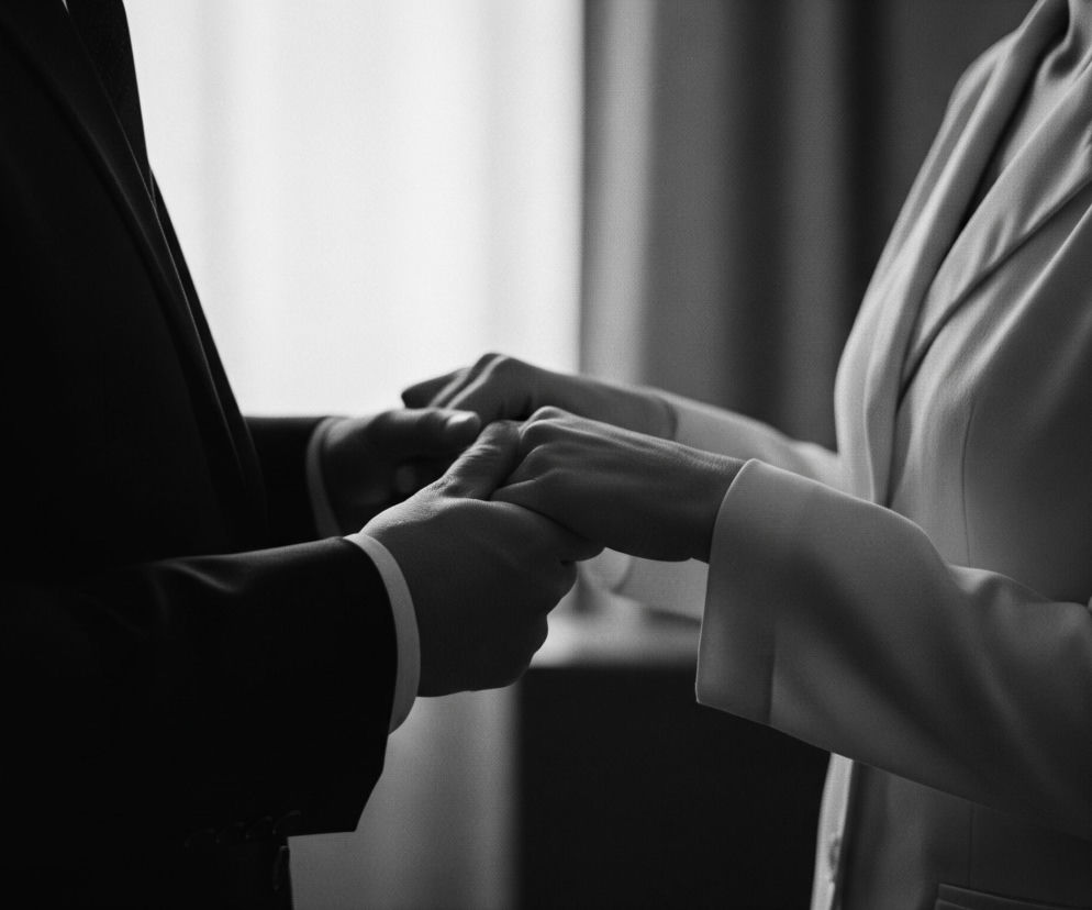 Na czarno-białym zdjęciu widać fragmenty dwóch osób, ujęte od ramion w dół, trzymających się za ręce. Po lewej stronie znajduje się mężczyzna w ciemnym garniturze i jasnej koszuli z mankietem, po prawej kobieta w jasnej marynarce z szerokimi klapami. Ich dłonie są splecione w delikatnym uścisku, przy czym dłonie kobiety obejmują dłonie mężczyzny. W tle widać niewyraźne, pionowe kształty, sugerujące zasłony lub ścianę, z jaśniejszym obszarem po lewej stronie, przypominającym okno.