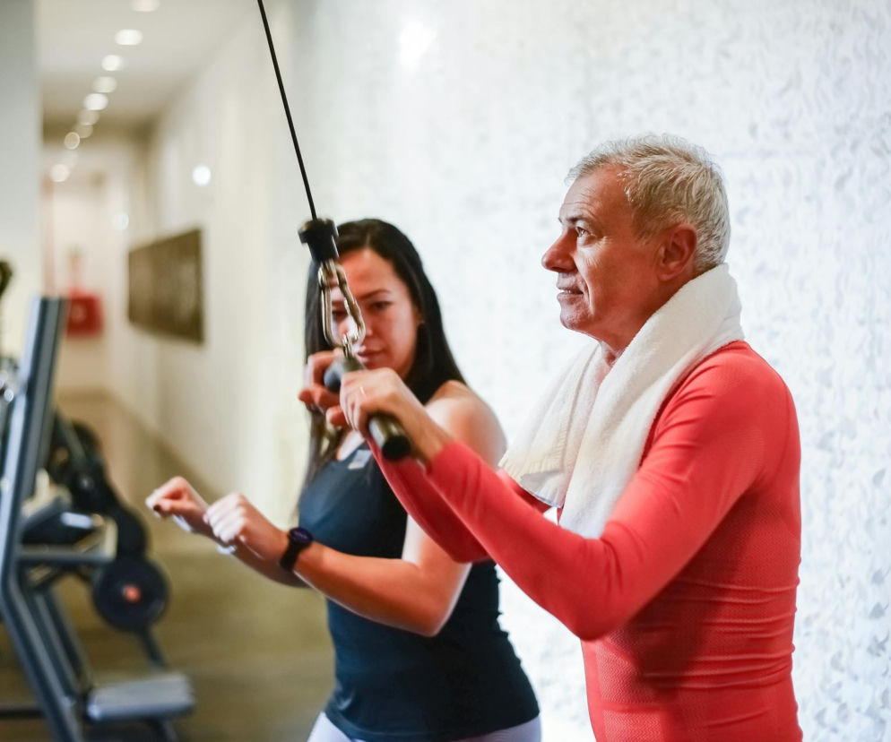 Starszy mężczyzna z ręcznikiem na szyi ćwiczy na maszynie do fizjoterapii pod okiem młodej kobiety, symbolizując wsparcie i rehabilitację w chorobie Parkinsona. Dowiedz się więcej o korzyściach fizjoterapii na Poradnik Zdrowie.