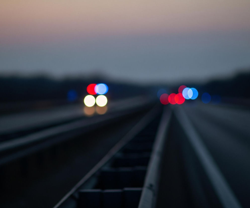 Rozmyta fotografia przedstawia autostradę o zmierzchu lub świcie. Na pierwszym planie widać bariery ochronne pośrodku drogi, składające się z kilku równoległych, ciemnych, podłużnych elementów. W tle, w oddali, znajdują się rozmyte światła samochodów: po lewej stronie cztery światła, dwa czerwone i dwa białe (lub żółte), a po prawej sześć świateł, cztery czerwone i dwa niebieskie. Horyzont jest rozmazany, z ciemnymi konturami drzew po bokach drogi, a niebo nad nimi przechodzi od jasnego, beżowo-różowego odcienia u góry do szaro-niebieskiego w niższej części.