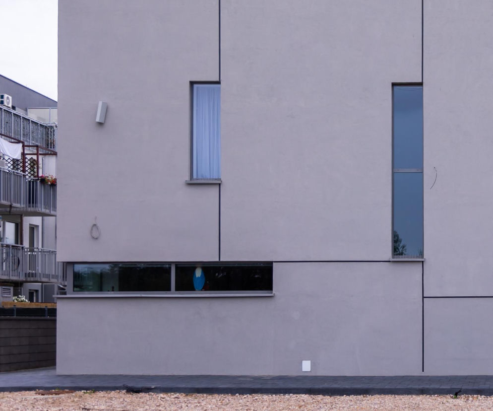 Fragment szarej fasady kościoła Matki Bożej Pompejańskiej na Białołęce z podłużnymi i poziomymi oknami, obok widoczne balkony sąsiedniego budynku. Obiekt, o którym można przeczytać na portalu Architektura Murator Plus, wyróżnia minimalistyczna forma architektoniczna.