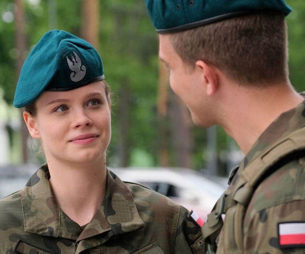 Młoda kobieta w mundurze wojskowym i zielonym berecie z orłem, uśmiecha się patrząc na mężczyznę w wojskowym stroju. Na mundurach widoczne polskie flagi, tło rozmyte. Scena odzwierciedla relacje z M jak miłość. Więcej o serialach na Super Seriale.