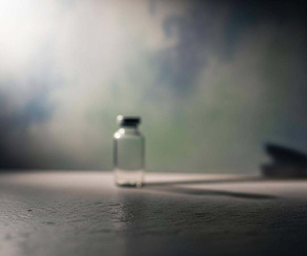 Na zdjęciu centralnie stoi mała, przezroczysta szklana fiolka z ciemną, cylindryczną nakrętką, rzucająca długi cień w prawo na jasnoszarym blacie. W tle widoczna jest rozmyta ściana z przejściami tonalnymi od jasnej bieli w lewym górnym rogu, przez zieleń i błękit, do ciemnego, nieokreślonego kształtu po prawej stronie. Cała scena jest utrzymana w stonowanych, chłodnych barwach, a oświetlenie tworzy dramatyczny kontrast między jasnymi i ciemnymi obszarami.