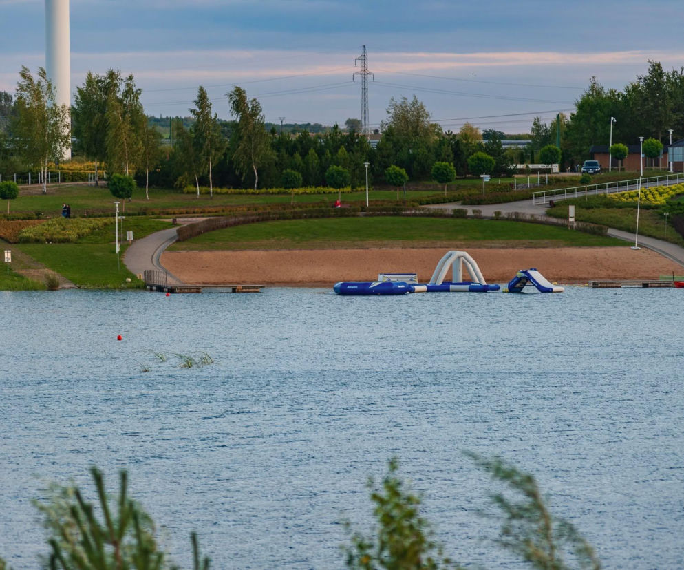 Widok na Kleczewską Maltę z wodnym placem zabaw na jeziorze, piaszczystą plażą i promenadą biegnącą wzdłuż brzegu, otoczoną zielenią oraz drzewami. W oddali widoczne turbiny wiatrowe. Więcej o rozbudowie przeczytasz na naszym portalu.