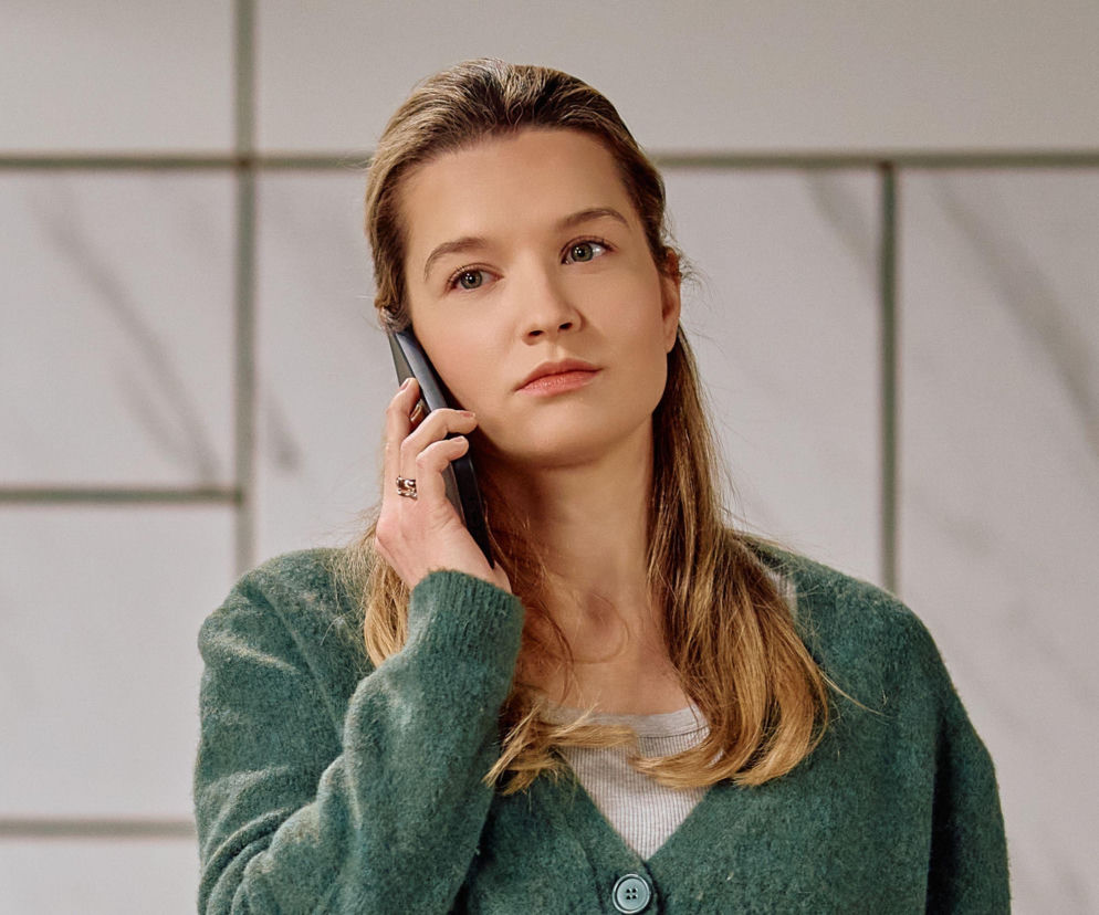 Kasia ze Na Wspólnej rozmawia przez telefon, wyglądając na zmartwioną lub zamyśloną. W tle widoczne są jasne, prostokątne kafelki. Więcej o planie Kasi wobec Kaliny przeczytasz na Super Seriale.