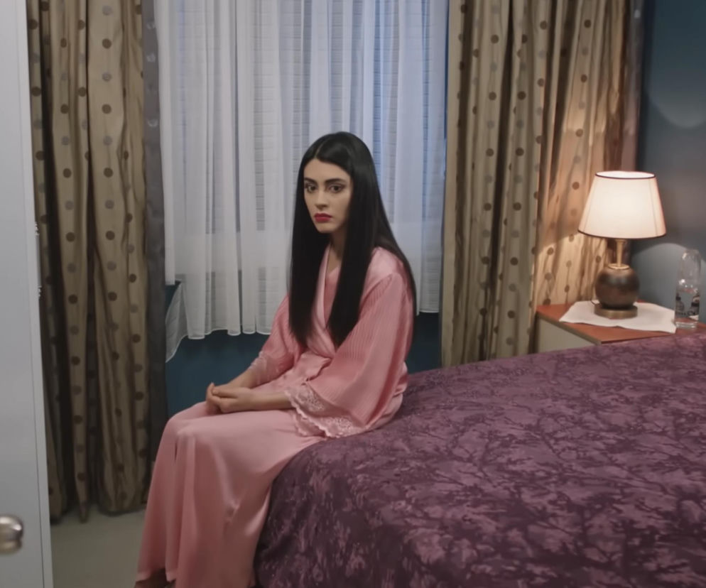 Hancer, młoda kobieta z długimi, ciemnymi włosami i czerwonymi ustami, siedzi na łóżku w sypialni, ubrana w różową koszulę nocną i peniuar. Jej zaniepokojona mina odzwierciedla intrygę Beyzy, która niszczy jej relację z mężem. Więcej o serialu Panna młoda i innych historiach miłosnych przeczytasz na Super Seriale.