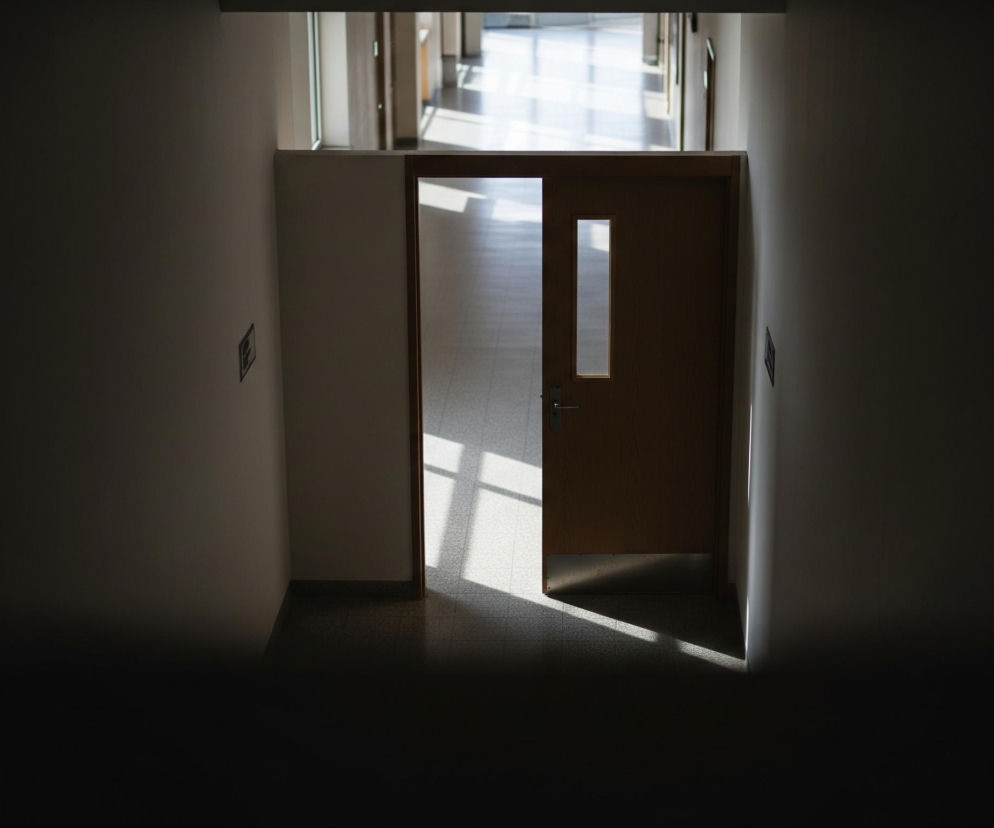 Wąski, ciemny korytarz prowadzi do jaśniejszej przestrzeni, widocznej za otwartymi drzwiami. Drewniane drzwi z pionowym, prostokątnym oknem pośrodku są osadzone w beżowej ścianie, na której po lewej stronie znajduje się ciemna, prostokątna tabliczka. Po prawej stronie, na podłodze z ciemnej, kratkowanej terakoty, leży jasny, trójkątny rzut światła słonecznego. Za drzwiami rozciąga się długi, szeroki korytarz, wyłożony jasnymi płytkami, gdzie również widoczne są intensywne pasma światła wpadającego przez okna po prawej stronie.