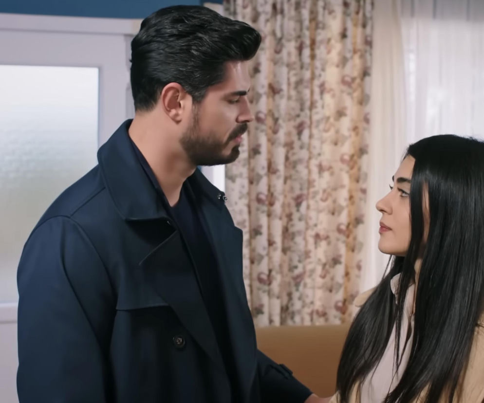 Kadr z serialu Panna młoda przedstawia Cihana w granatowym płaszczu i Hancer w beżowym, spoglądającą na niego. Scena rozgrywa się w pomieszczeniu z jasnymi drzwiami i zasłonami w kwiatowy wzór, symbolizując napięcie w ich relacji, o której przeczytasz na Super Seriale.