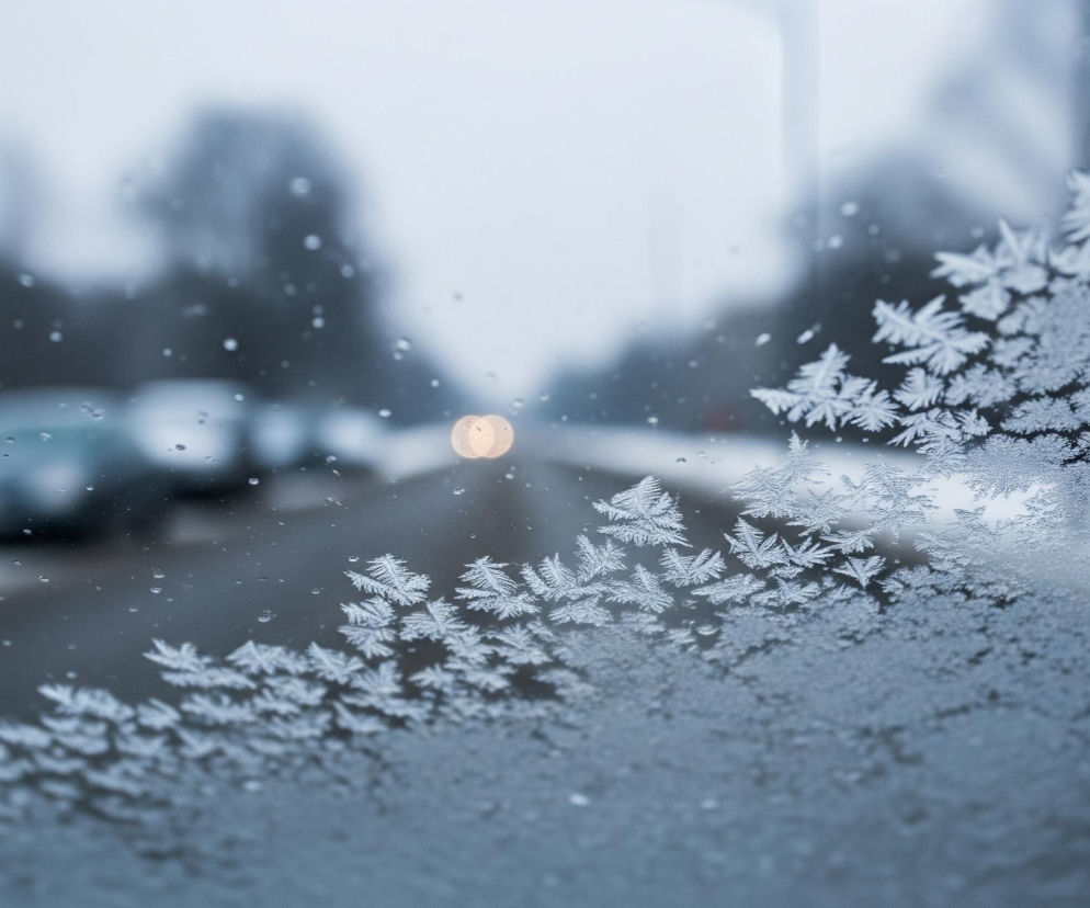 Prawa część przedniej szyby pokryta jest warstwą szronu w kształcie rozgałęzionych, przypominających paprocie wzorów, które są wyraźnie widoczne i ostro zarysowane. Lewa część szyby jest częściowo mokra, z widocznymi pojedynczymi kroplami wody, a szron jest mniej gęsty i bardziej rozproszony. Za szybą rozciąga się miejska ulica, oświetlona rozmytym, pomarańczowym światłem, z zaparkowanymi samochodami po lewej stronie drogi. Tło jest zamglone, dominują szare i niebieskawe odcienie, co sugeruje zimowy, pochmurny dzień.