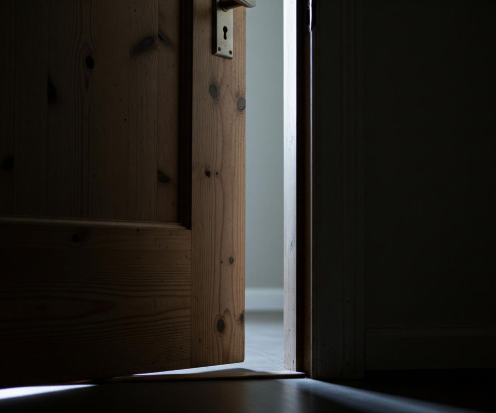 Otworzone, drewniane drzwi w kolorze jasnego brązu widoczne są po lewej stronie kadru, z mosiężną klamką i zamkiem umieszczonymi w górnej części. Na powierzchni drzwi widać wyraźne słoje drewna oraz ciemniejsze sęki. Pomiędzy drzwiami a futryną widać wąski pasek jasnego światła, które delikatnie oświetla podłogę pod drzwiami. Reszta pomieszczenia po prawej stronie drzwi jest pogrążona w ciemności, tworząc kontrast z jasnym prześwitem.