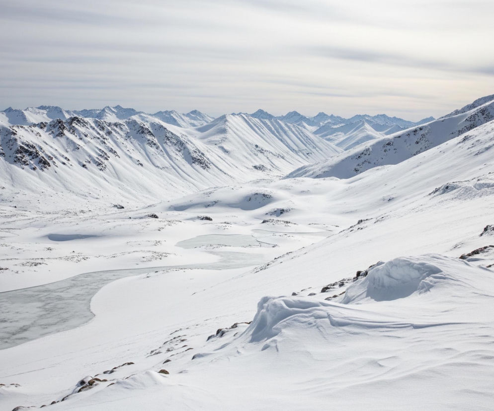 Rozległa, zimowa panorama przedstawia pasmo górskie i dolinę, pokryte grubą warstwą śniegu. W tle rozciągają się ośnieżone szczyty górskie o ostrych zboczach, a na pierwszym planie po lewej stronie znajduje się zamarznięty zbiornik wodny w kształcie nieregularnej, wydłużonej rzeki. Dolina wypełniona jest śniegiem, z wystającymi miejscami ciemnymi plamami skał lub roślinności, zwłaszcza na zboczach gór oraz w dolnej części obrazu. Niebo nad górami jest jasne, z widocznymi poziomymi warstwami chmur.
