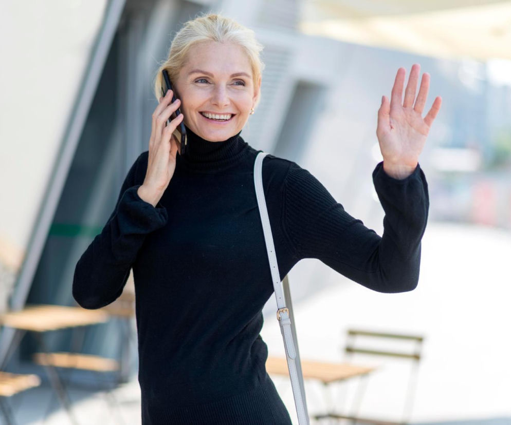 Blondynka w czarnym golfie, uśmiechnięta, rozmawia przez telefon i macha, stojąc na zewnątrz. Symbolizuje nowoczesne sposoby utrzymywania kontaktu z bliskimi na odległość, o których przeczytasz więcej na naszym portalu.