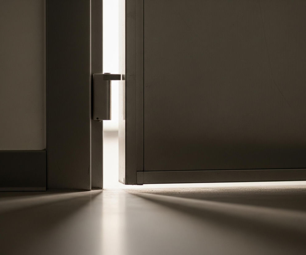 Częściowo otwarte drzwi, znajdujące się w centralnej części kadru, ukazują pionową smugę jasnego światła pomiędzy ciemnymi panelami ościeżnicy i skrzydła. Skrzydło drzwi jest ciemnoszare i nieco szorstkie, z widoczną krawędzią po prawej stronie. Na panelu po lewej stronie znajduje się metalowy, srebrny lub jasnoszary element zamka. Od jasnego paska światła padają promienie, tworząc długie, rozchodzące się cienie na podłodze, która jest nieco jaśniejsza i gładka. Górna część obrazu i większość bocznych fragmentów toną w ciemności, podkreślając kontrast z centralnym, świetlistym elementem.