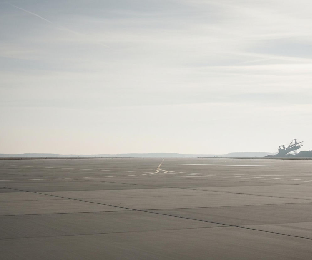 Szeroki, asfaltowy pas startowy, w odcieniach szarości, zajmuje dolną połowę kadru, rozciągając się w kierunku horyzontu. Widoczne są jasne, białe i żółte linie poziome i pionowe, wyznaczające jego powierzchnię. Na odległym horyzoncie, w prawym fragmencie, zza niskiego, ciemnego wzniesienia wyłania się duża, postindustrialna konstrukcja przypominająca koparkę wielonaczyniową, jej ciemna sylwetka jest niewyraźna na tle zamglonego, jasnoszarego nieba z nielicznymi, cienkimi smugami chmur.