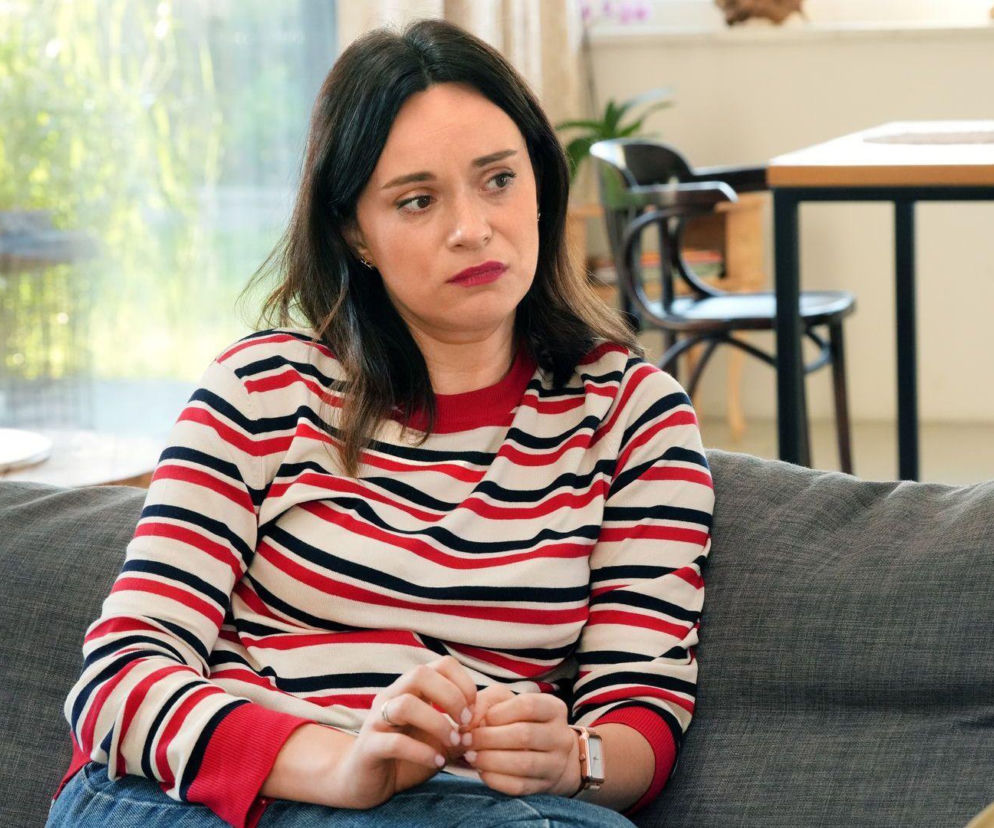 Kobieta z ciemnymi włosami w czerwono-granatowo-beżowym swetrze w paski, siedząca na szarej kanapie ze splecionymi dłońmi, ze zmartwioną miną, w tle rozmazane elementy wnętrza. Zdjęcie przedstawia aktorkę Marię Dejmek, serialową Natalię z Barw szczęścia. Więcej o tym na Super Seriale.