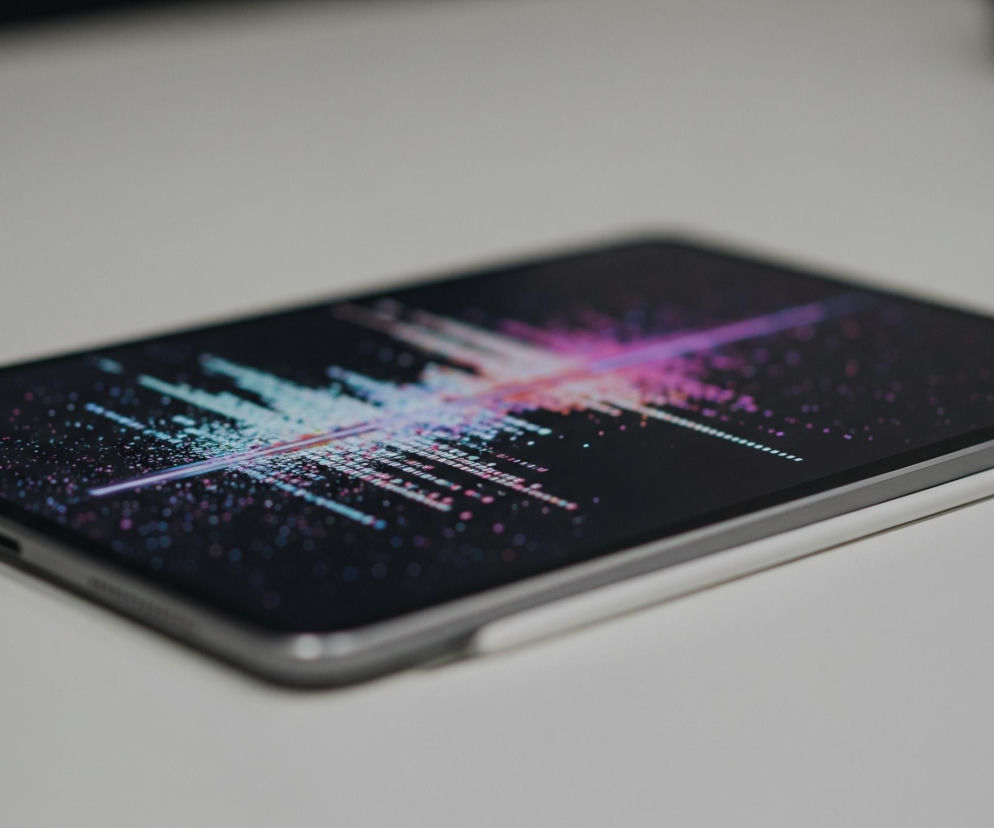 Na białej powierzchni leży tablet z ciemnym ekranem skierowanym w górę i lekko pod kątem. Ekran wyświetla abstrakcyjny wzór składający się z drobnych, kolorowych punktów i linii w odcieniach turkusu, fioletu i różu, które promieniują od centrum w lewo i prawo. Do prawej krawędzi tabletu, magnetycznie przymocowany jest biały rysik.