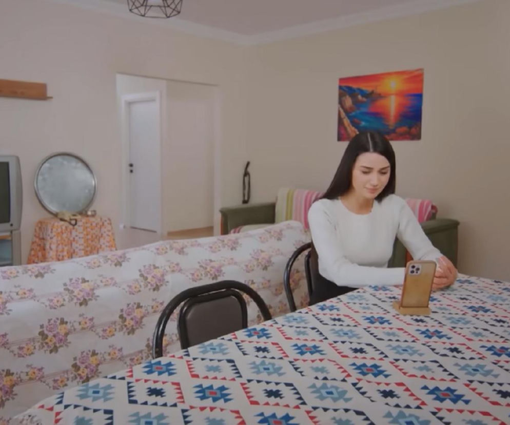 Bahar z serialu Miłość i nadzieja nagrywa wideo w skromnym mieszkaniu, prezentując się jako ofiara, aby oczernić Kuzeya. Widoczna kobieta z długimi ciemnymi włosami w białym swetrze, używająca smartfona do nagrania. Scena przedstawia jej plan zemsty, który można śledzić na Super Seriale.