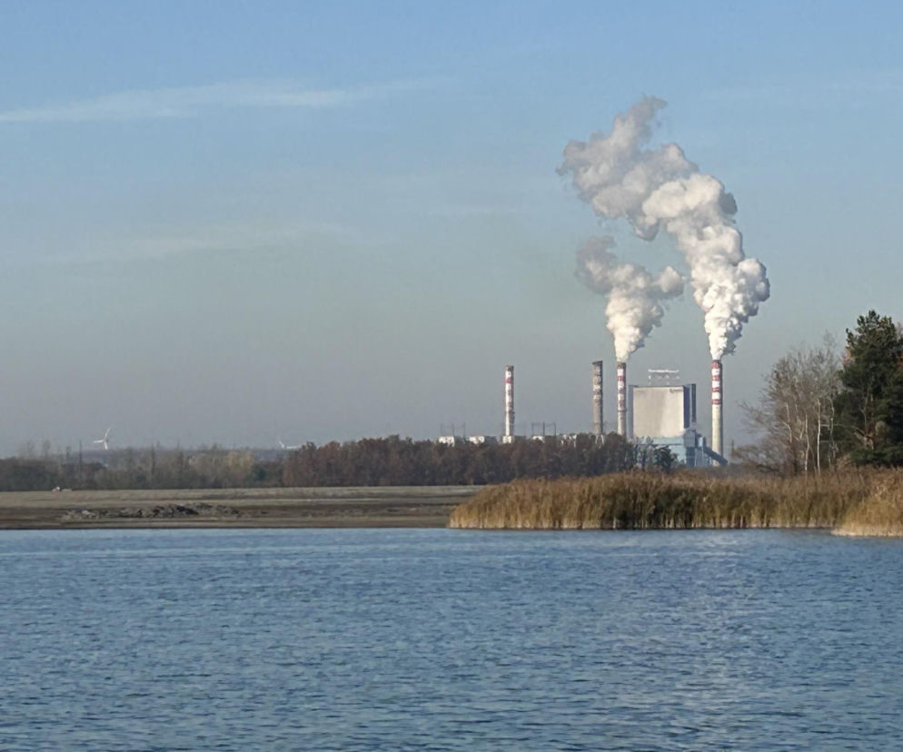 Dymiące kominy elektrowni Pątnów z widocznymi turbinami wiatrowymi na horyzoncie, odbijające się w błękitnej tafli wody jeziora. Trzciny na brzegu dopełniają krajobraz, symbolizujący przemiany energetyczne w regionie, o których przeczytasz na naszym portalu.