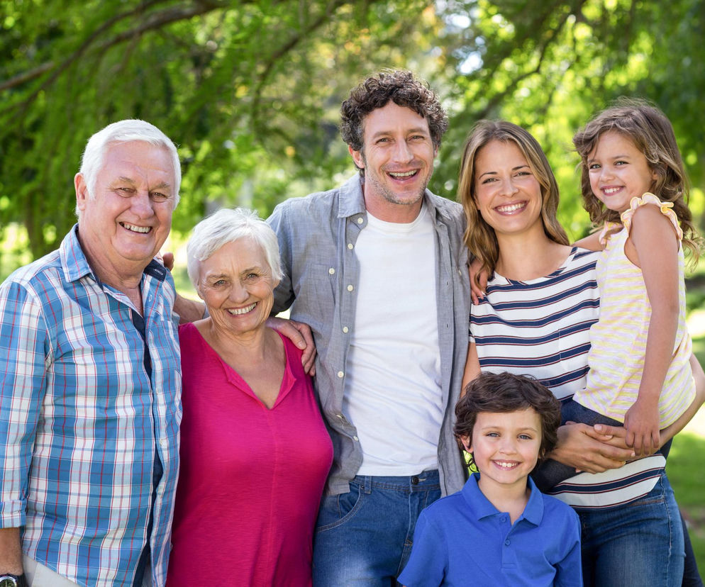 Uśmiechnięta rodzina – dziadkowie, rodzice i dwoje dzieci, stojąca w parku. Seniorzy, którzy wychowali dzieci i nie otrzymali wsparcia, domagają się wstecznego 800 plus. Więcej o tym, czy emeryci dostaną pieniądze za wychowanie dzieci, przeczytasz na Poradnik Zdrowie.
