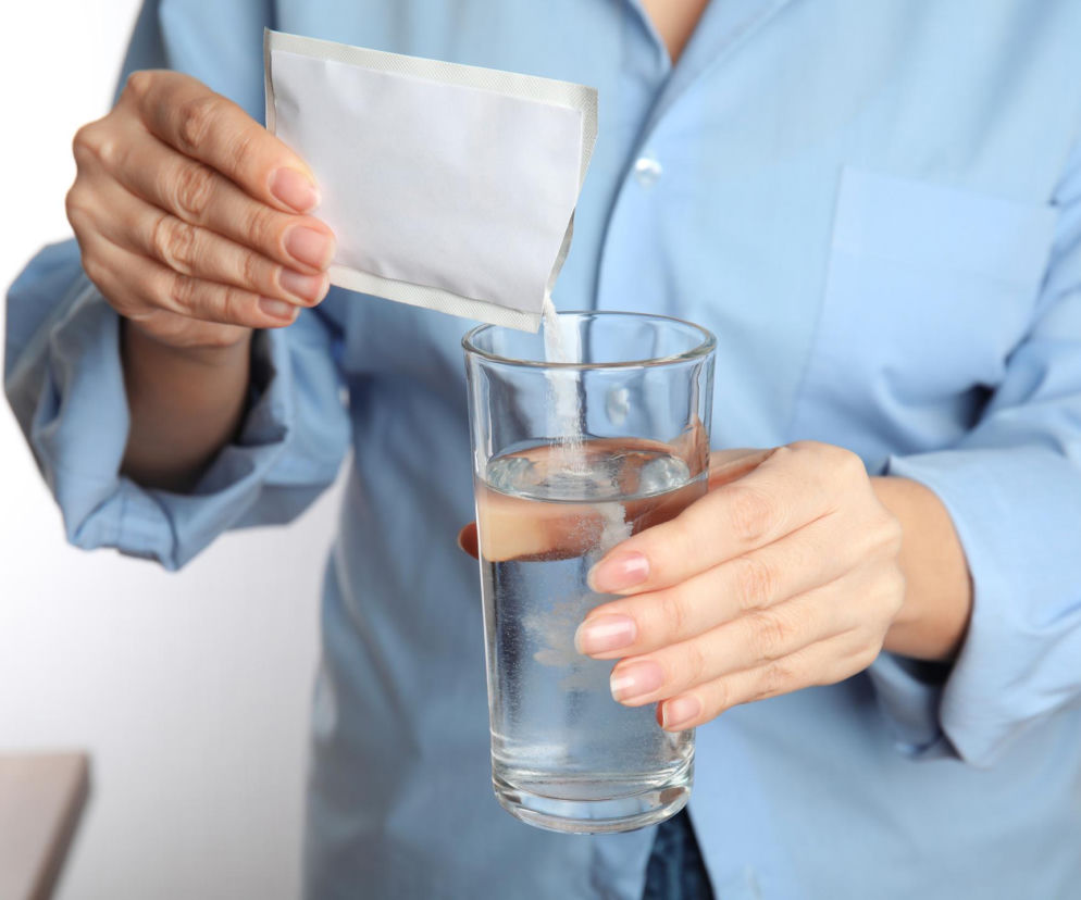Kobieta w błękitnej koszuli wsypuje biały proszek z saszetki do szklanki z wodą. Zdjęcie symbolizuje spożywanie suplementów diety, co w kontekście artykułu na Poradnik Zdrowie przestrzega przed skażonymi produktami.