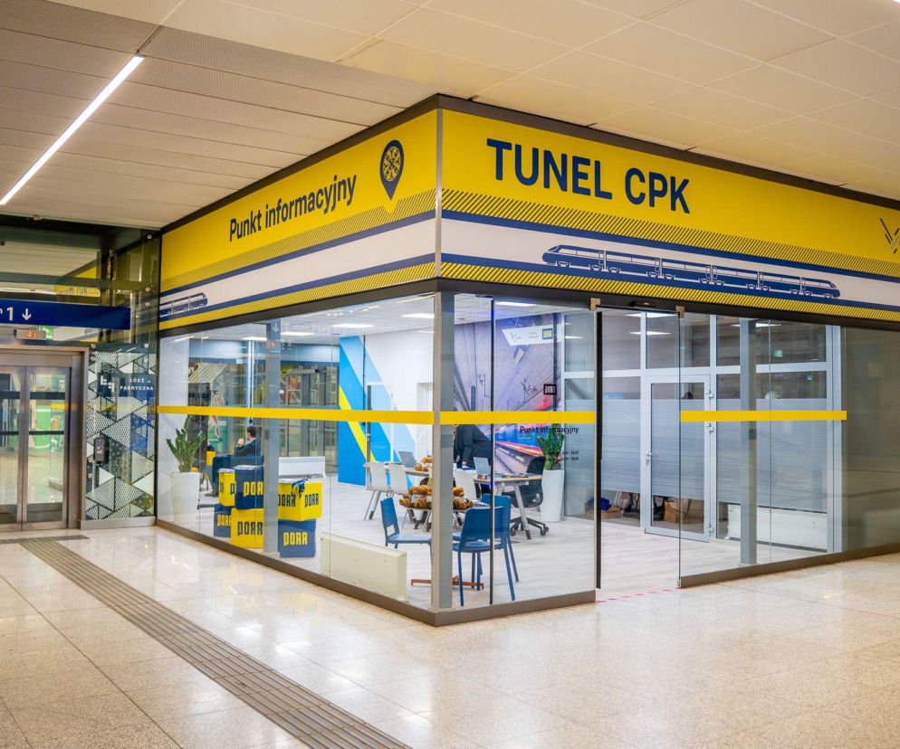 Punkt informacyjny Tunel CPK na dworcu Łódź Fabryczna w żółto-niebieskiej kolorystyce, z widocznym logo PORR i wizualizacją kolei dużych prędkości, stanowiący centrum komunikacji z mieszkańcami o projekcie tunelu, o którym można przeczytać więcej na Architektura Murator Plus.