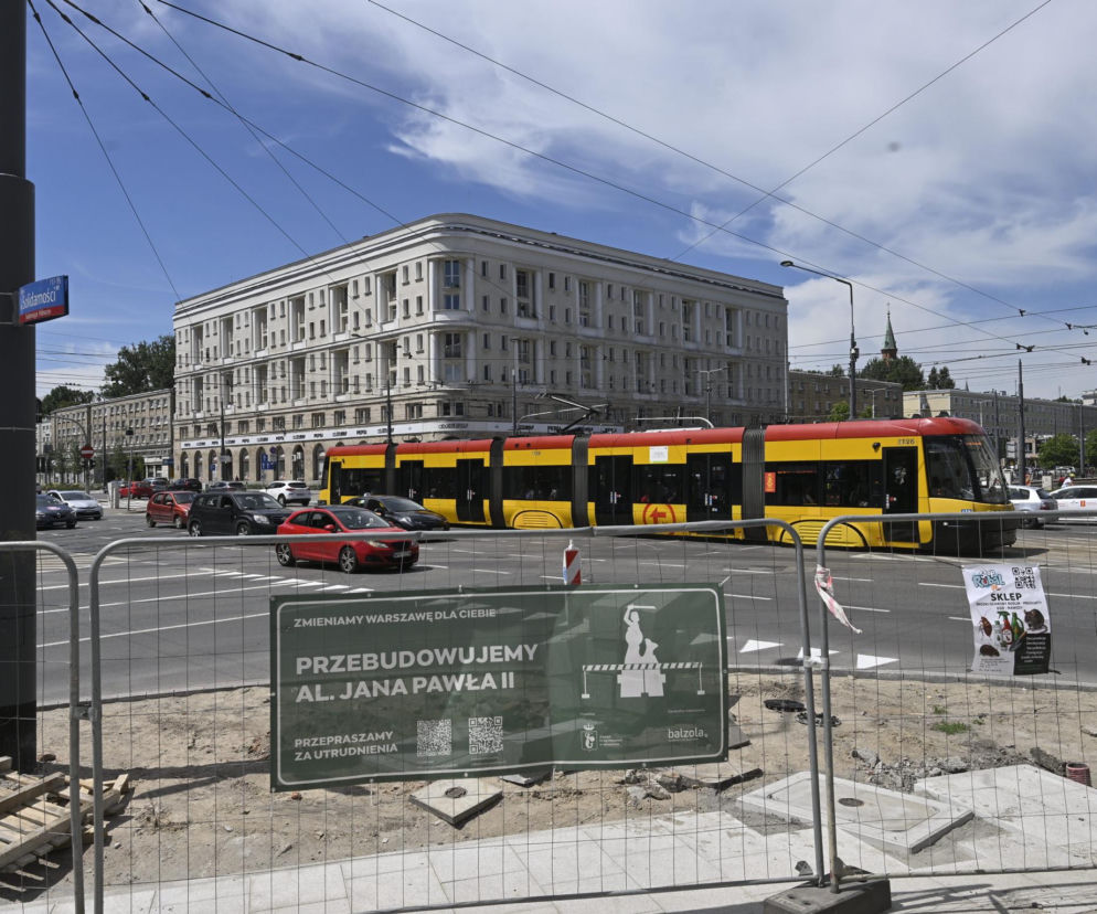 Tramwaj czerwono-żółty jedzie przez skrzyżowanie Al. Solidarności i Jana Pawła II w Warszawie. Obok torów widać plac budowy z siatką ogrodzeniową i tablicę informującą o przebudowie Al. Jana Pawła II. Więcej o remontach torowisk przeczytasz na naszym portalu.