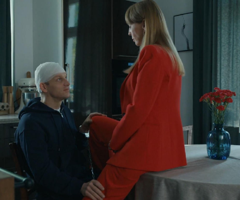 Agnieszka (Katarzyna Tlałka) w czerwonym garniturze siedząca na stole, patrzy na Łukasza (Michał Rolnicki) z opatrunkiem na głowie, który ma skrępowane nogi łańcuchem. Para aktorów w scenie z serialu Barwy szczęścia, której opis znajdziesz na Super Seriale.