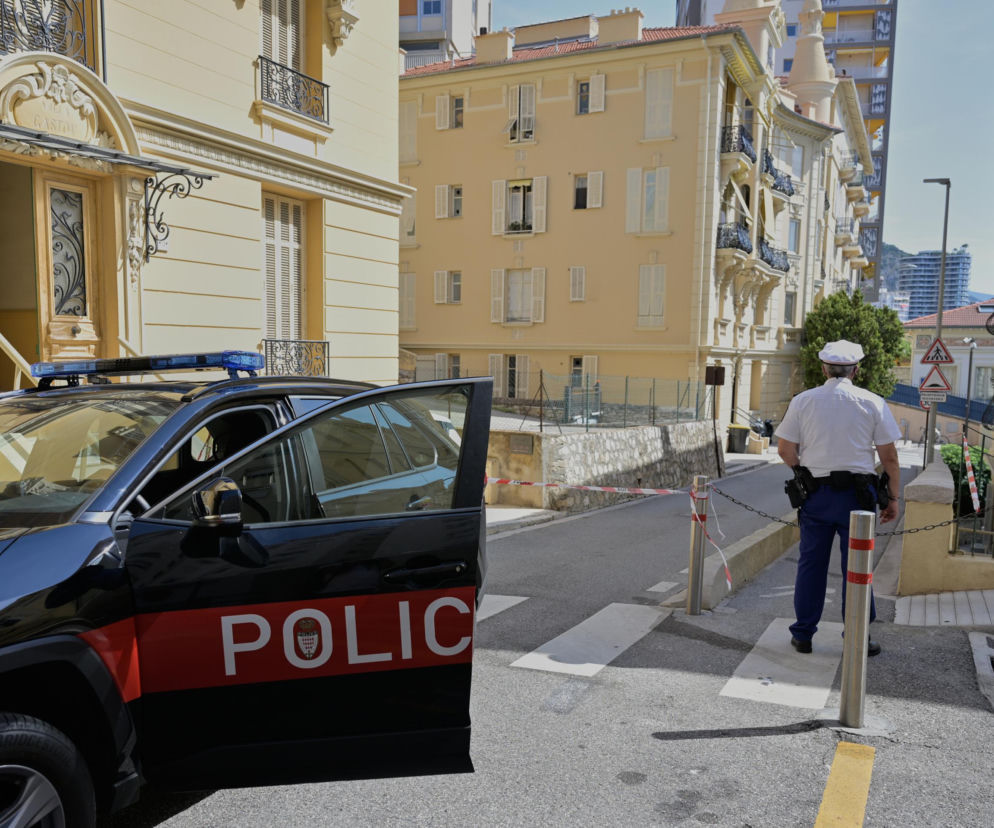 Radiowóz policji w Monako z otwartymi drzwiami i funkcjonariuszem patrolującym ulicę, częściowo wygrodzoną biało-czerwoną taśmą. Budynki w tle tworzą tło śledztwa dotyczącego eksplozji, o której przeczytasz więcej na naszym portalu.