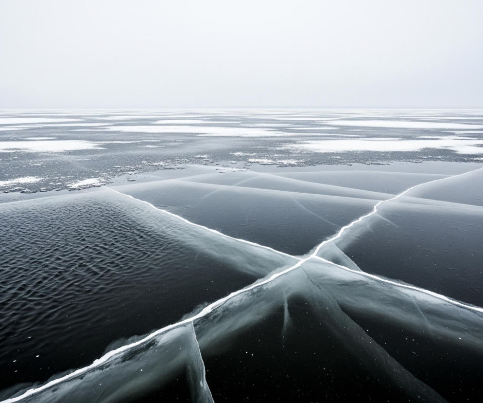 Zamarznięta powierzchnia wody z wyraźnymi pęknięciami i fragmentami lodu rozciąga się w nieskończoność pod mglistym, jasnym niebem. Na pierwszym planie dominują ciemnoniebieskie i szare odcienie lodu, poprzecinane jaśniejszymi, niemal białymi liniami pęknięć, które tworzą nieregularny wzór przecinających się krzyży. Po lewej stronie lód jest pofałdowany, przypominając drobne fale, natomiast po prawej i w centrum widoczne są gładkie, przezroczyste fragmenty, przez które przebijają się ciemniejsze barwy. Horyzont jest niewyraźny, zlewając się z mlecznym niebem, które zajmuje górną połowę obrazu.