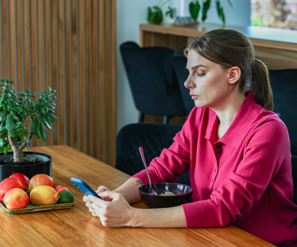 Kobieta w różowej bluzce siedząca przy drewnianym stole, patrząca w ekran smartfona. Przed nią talerz z owocami i miska. Zdjęcie ilustruje codzienność bohaterów, o których można przeczytać na Super Seriale.