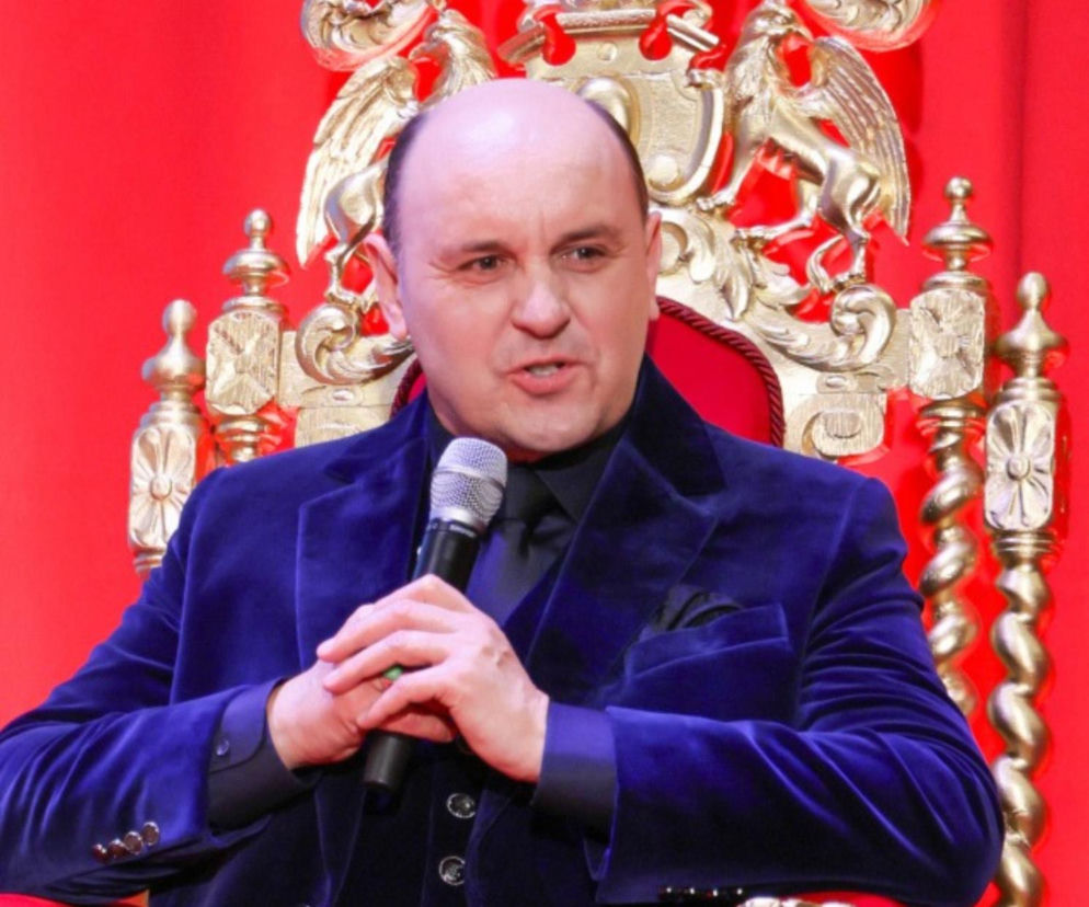 Adam Woronowicz w niebieskiej marynarce trzyma mikrofon, siedząc na złotym tronie. O show Taskmaster przeczytasz na Eska.