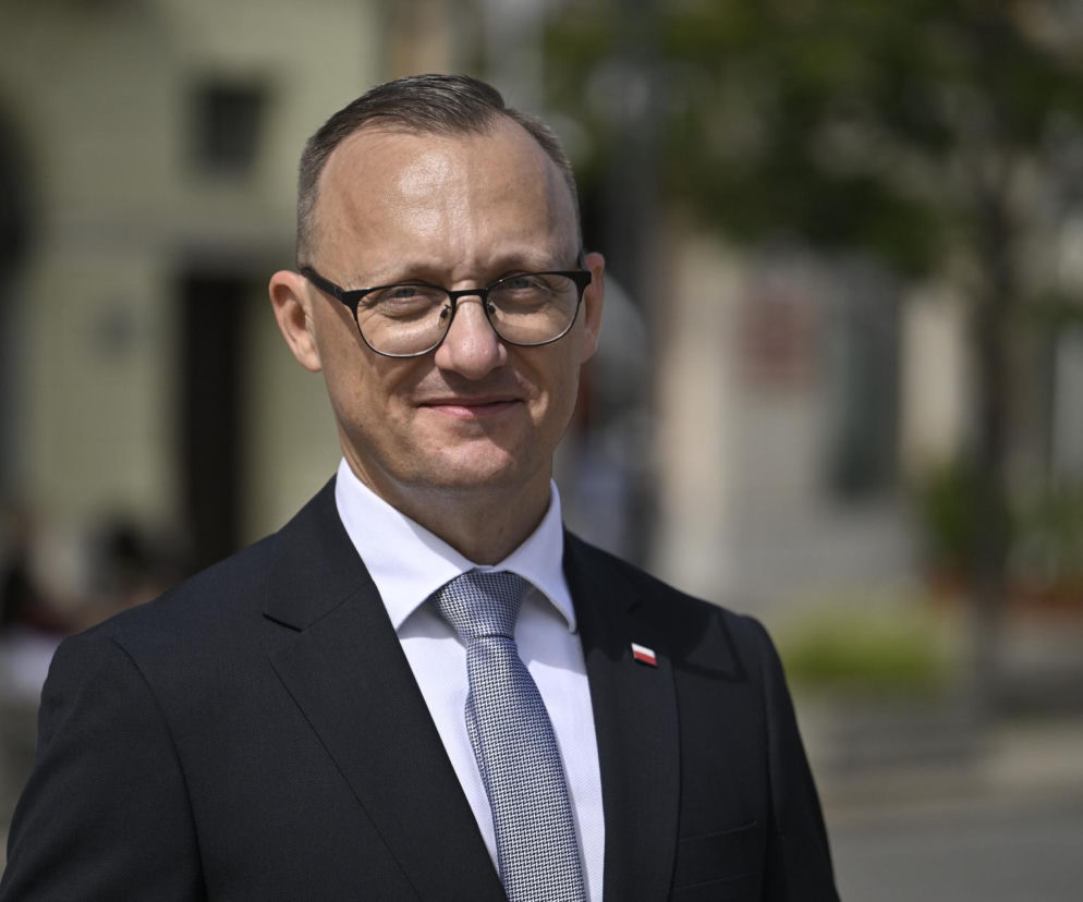 Rzecznik prezydenta Rafał Leśkiewicz w garniturze i krawacie, z przypinką polskiej flagi na marynarce, uśmiecha się lekko do kamery, co można przeczytać na naszym portalu. Mężczyzna stoi na zewnątrz w jasnym otoczeniu, rozmyte tło z budynkami i zielenią.
