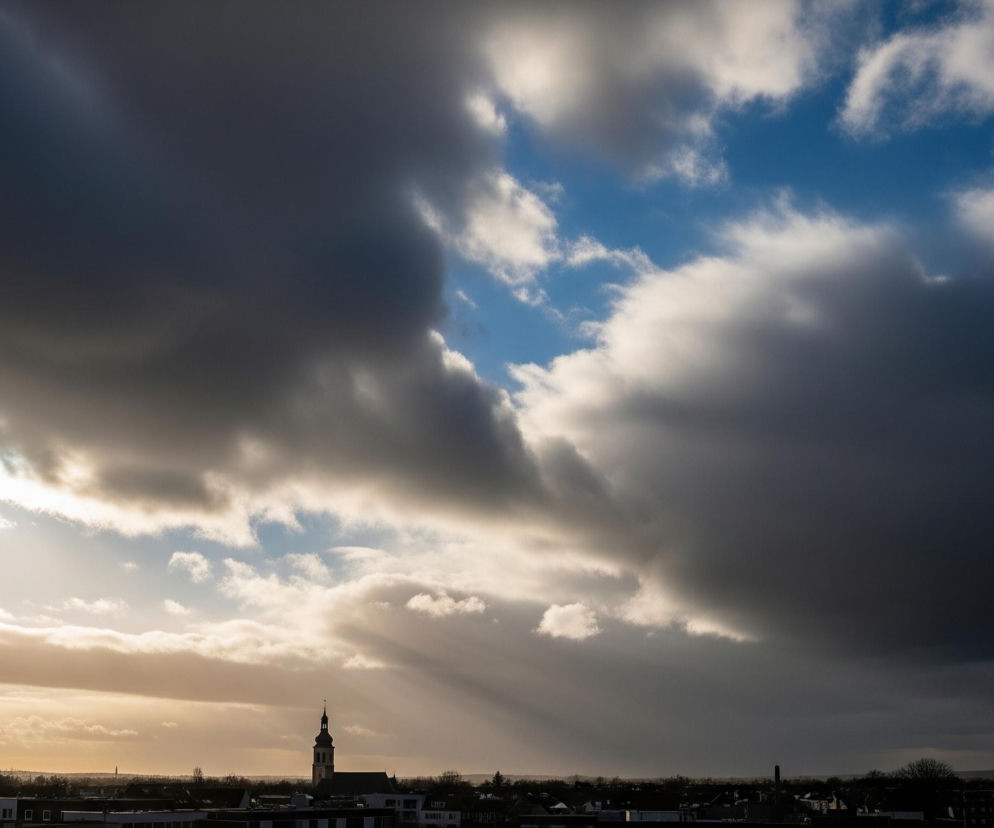 Niebo zdominowane jest przez dynamiczne chmury, częściowo ciemne i burzowe po prawej stronie, a częściowo jaśniejsze, przepuszczające promienie słońca po lewej i w centralnej części obrazu. U góry po prawej widoczny jest fragment czystego, błękitnego nieba. Horyzont zarysowany jest przez sylwetki budynków i drzew, a pośrodku kadru wyróżnia się wieża kościoła z ostro zakończonym dachem. Słońce, ukryte za chmurami po lewej stronie, rzuca jasne, złociste światło, tworząc smugi promieni na niebie i oświetlając fragmenty pejzażu.