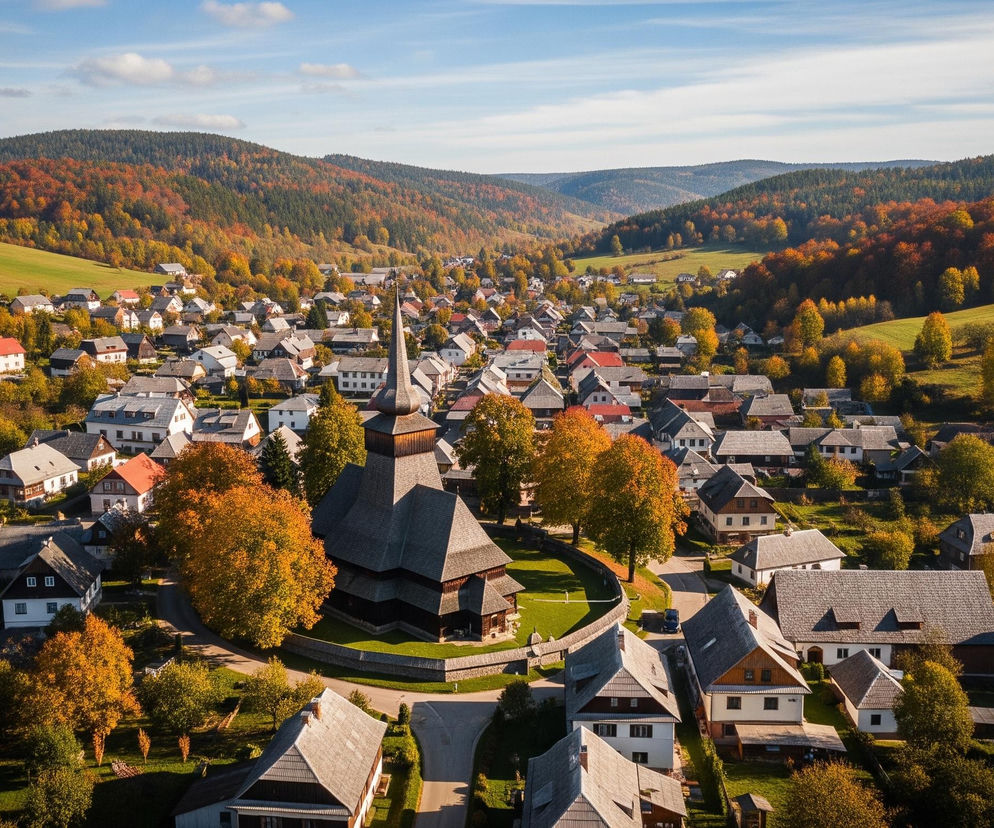 Centralnym punktem obrazu jest drewniany kościół o ciemnych ścianach i spiczastym dachu, otoczony gęstymi drzewami o jaskrawożółtych i pomarańczowych liściach. Wokół kościoła i rozciągających się w dolinie pagórkowatych zboczach rozmieszczone są liczne domy z białymi fasadami i ciemnymi dachami, niektóre z czerwonymi akcentami. Cały krajobraz jest usiany zielonymi polami i gęstymi lasami, które na horyzoncie przybierają ciemniejszy, jesienny odcień, wszystko to pod częściowo zachmurzonym, błękitnym niebem.
