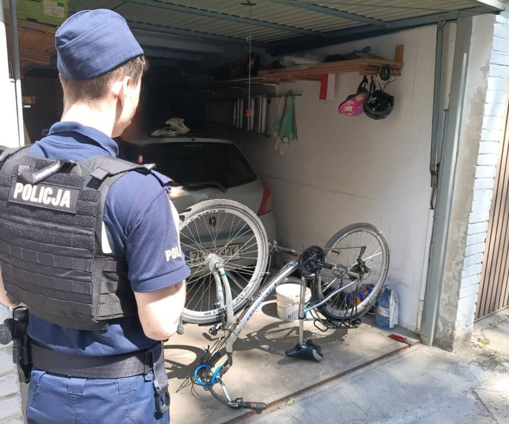 Policjant w mundurze i kamizelce taktycznej z napisem POLICJA stoi tyłem, obserwując garaż z zaparkowanym autem i rowerem leżącym na boku. Zdjęcie ilustruje działania profilaktyczne koszalińskiej policji w zakresie znakowania i zabezpieczeń rowerów, o czym więcej przeczytasz na naszym portalu.