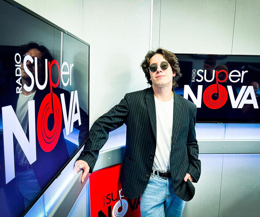 Dawid Kwiatkowski w okularach przeciwsłonecznych, ubrany w marynarkę w paski i jasne jeansy, opiera się o ekran z logo Radia SuperNova. Artysta publicznie opowiadał o swojej walce z boreliozą, o której więcej przeczytasz na Poradnik Zdrowie.