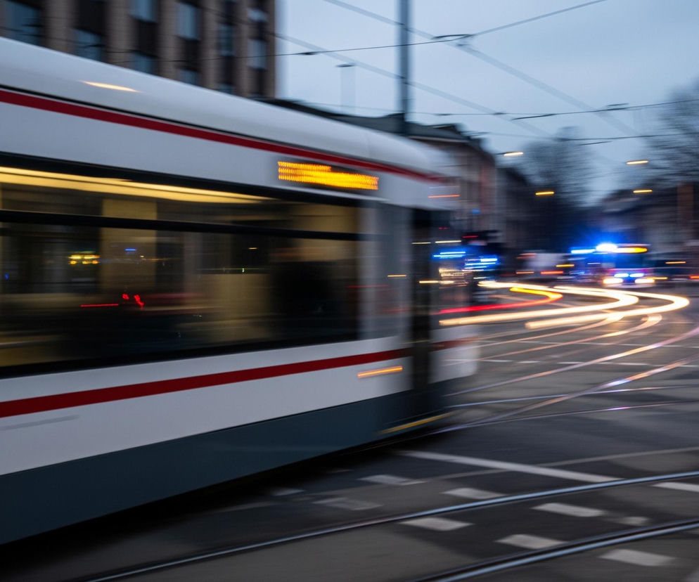 Szybko jadący biały tramwaj z czerwonym pasem biegnącym wzdłuż jego boku dominuje w lewej części kadru. Rozmyte światła uliczne i budynki tworzą tło, podczas gdy na drodze widać długie smugi świateł pojazdów – żółte z przodu i czerwone z tyłu. Na drugim planie, wśród rozmytych świateł, wyróżniają się niebieskie migające światła alarmowe, a tuż za nimi, z prawej strony kadru, widać samochód z włączonymi światłami, stojący na skrzyżowaniu torów tramwajowych. Całość kompozycji, wykonana z długim czasem naświetlania, oddaje dynamikę ruchu miejskiego o zmierzchu lub w nocy.