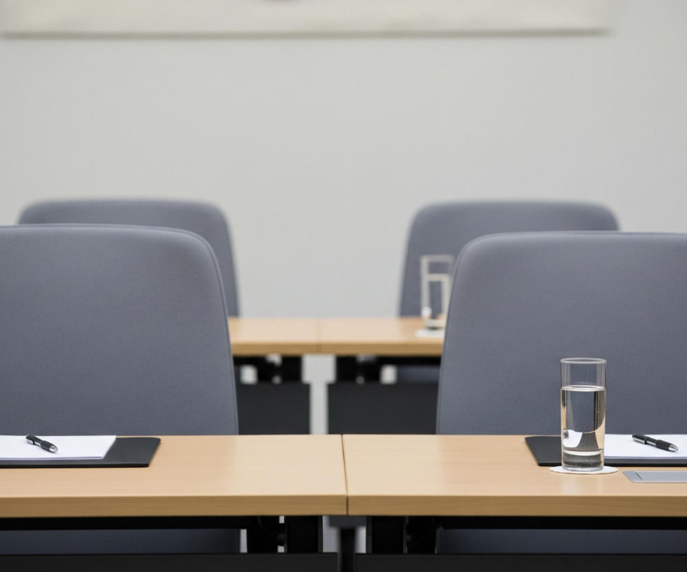 Na pierwszym planie widoczne są dwa rzędy drewnianych stołów konferencyjnych z szarymi krzesłami. Na każdym stole, przed krzesłem, znajduje się wysoka szklanka napełniona do połowy wodą, umieszczona na białej podkładce, obok której leży czarny notatnik z białą kartką i czarny długopis. Tło stanowi jasnoszara ściana, a dalsze rzędy stołów i krzeseł są rozmyte.