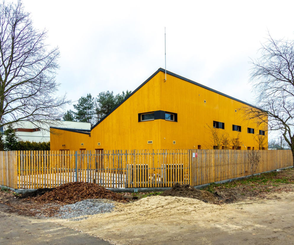 Dom Młodej Matki w Warszawie, projekt biura WXCA, z żółtą elewacją z drewna i spadzistym dachem, otoczony drewnianym płotem. Obiekt oferuje bezpieczne środowisko dla nastoletnich matek i ich dzieci. Więcej o architekturze tego typu przeczytasz na Architektura Murator Plus.